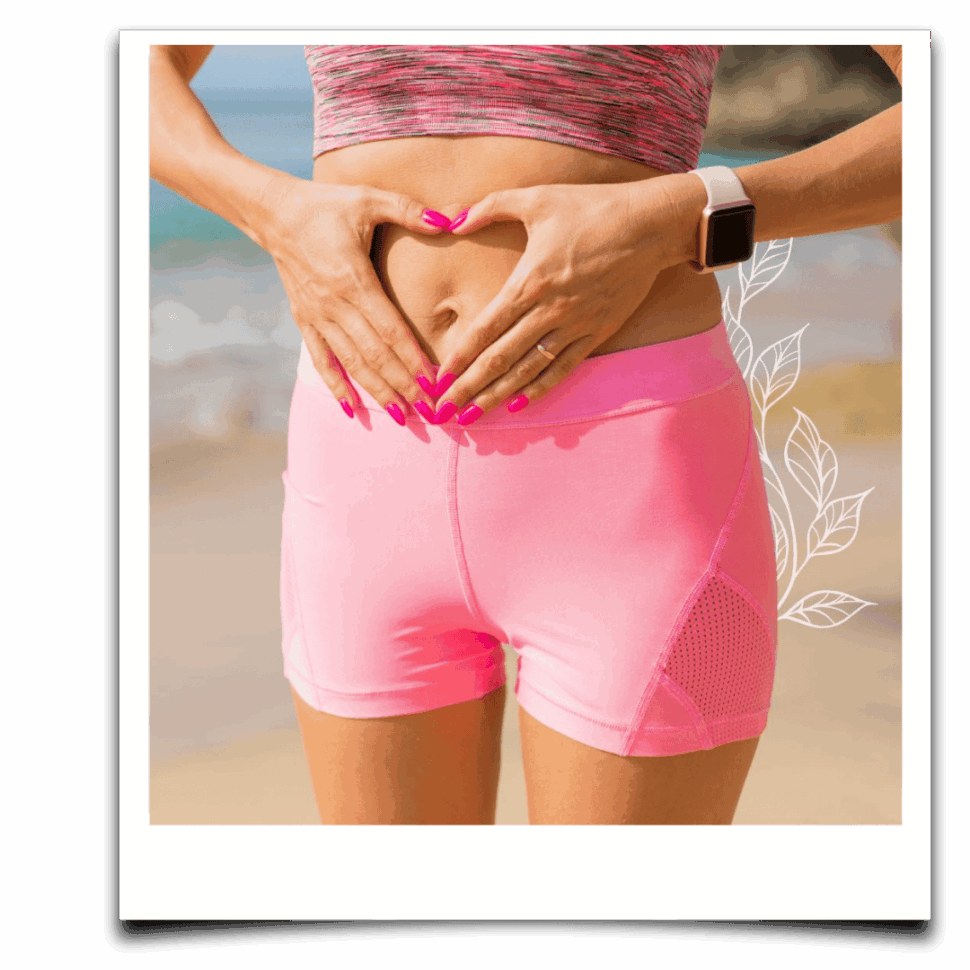 Žena ve sportovním oblečení drží ruce ve tvaru srdce na oblasti břicha u moře, jako gesto péče o trávení.