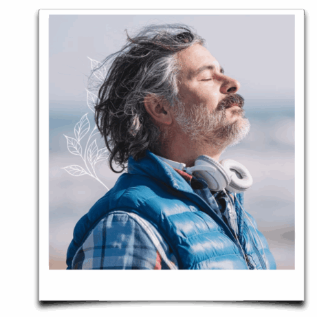 Starší muž se sluchátky kolem krku tiše dýchá u moře se zavřenýma očima – vyzařuje klid, propojení a vnitřní vyrovnanost.