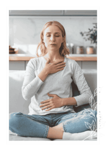 Žena sedí v tureckém sedu na gauči, má zavřené oči a obě ruce položené na hrudi a břiše – vědomě dýchá a zklidňuje se.